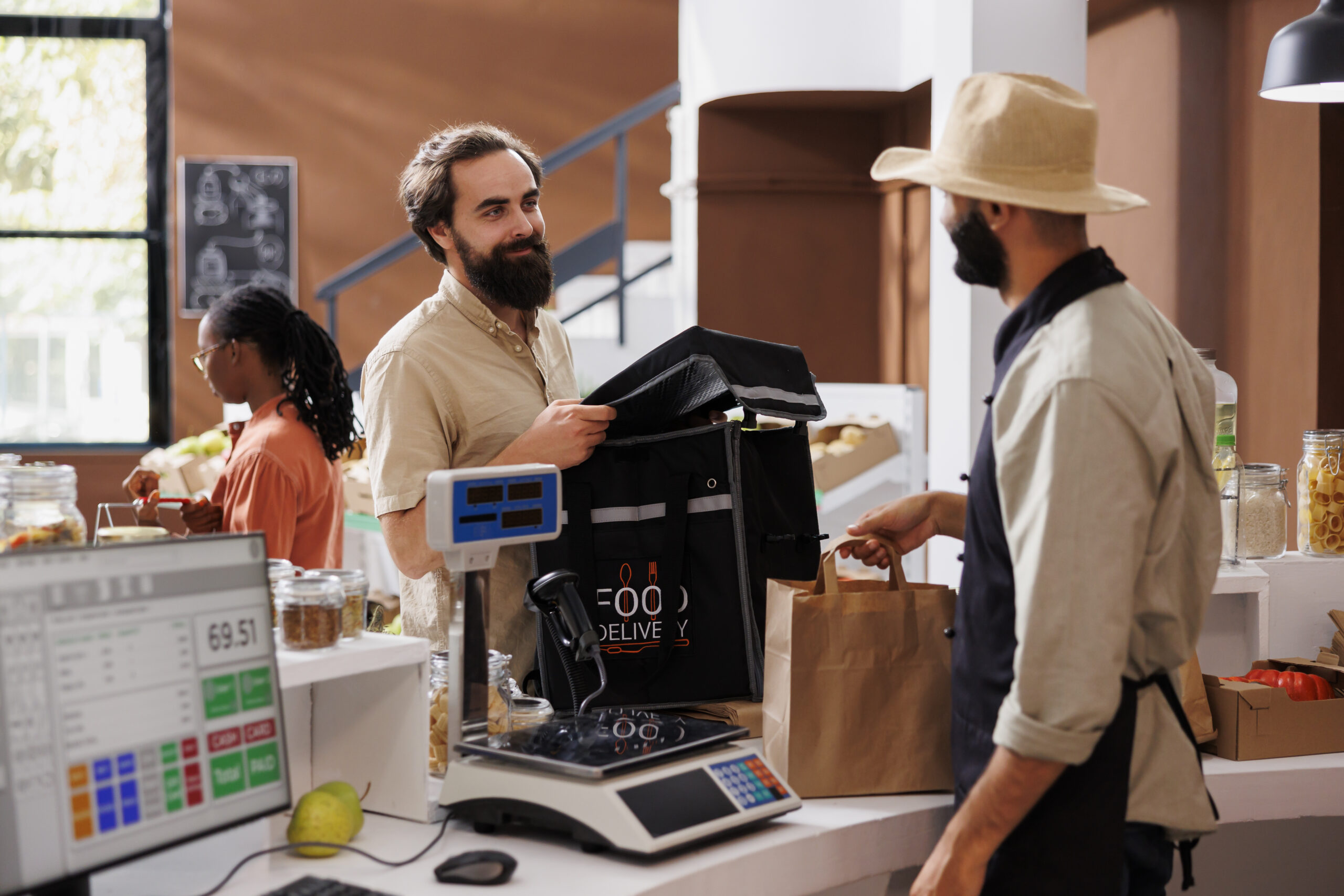 male courier arrives at checkout counter ready to receive packaged grocery order from cashier. Food delivery man with backpack retrieves takeaway from vendor at local market.